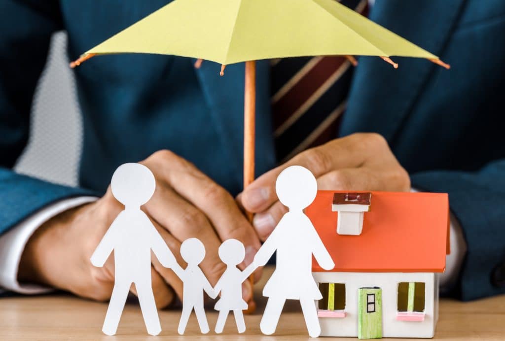An insurance agent holds a toy umbrella over a paper cutout family and house figurine.